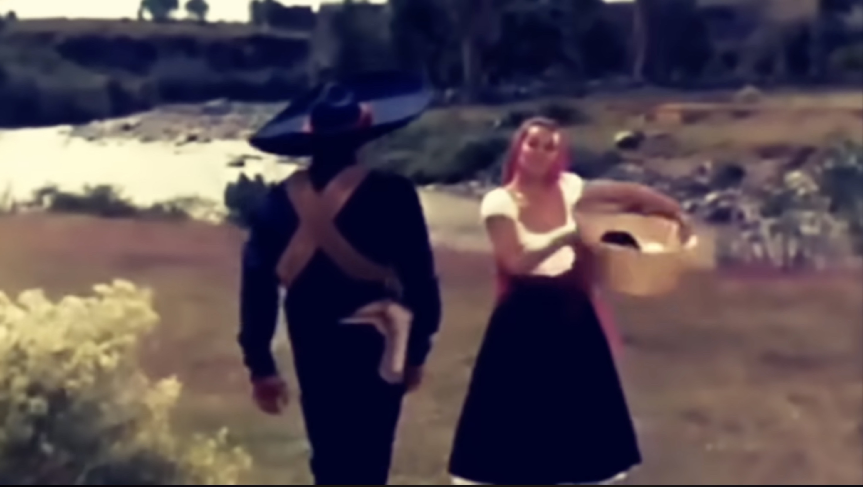 Still from La Cucaracha (1959) showing a soldier in black with a crossed bandolier walking beside a woman in lighter clothing carrying a basket.