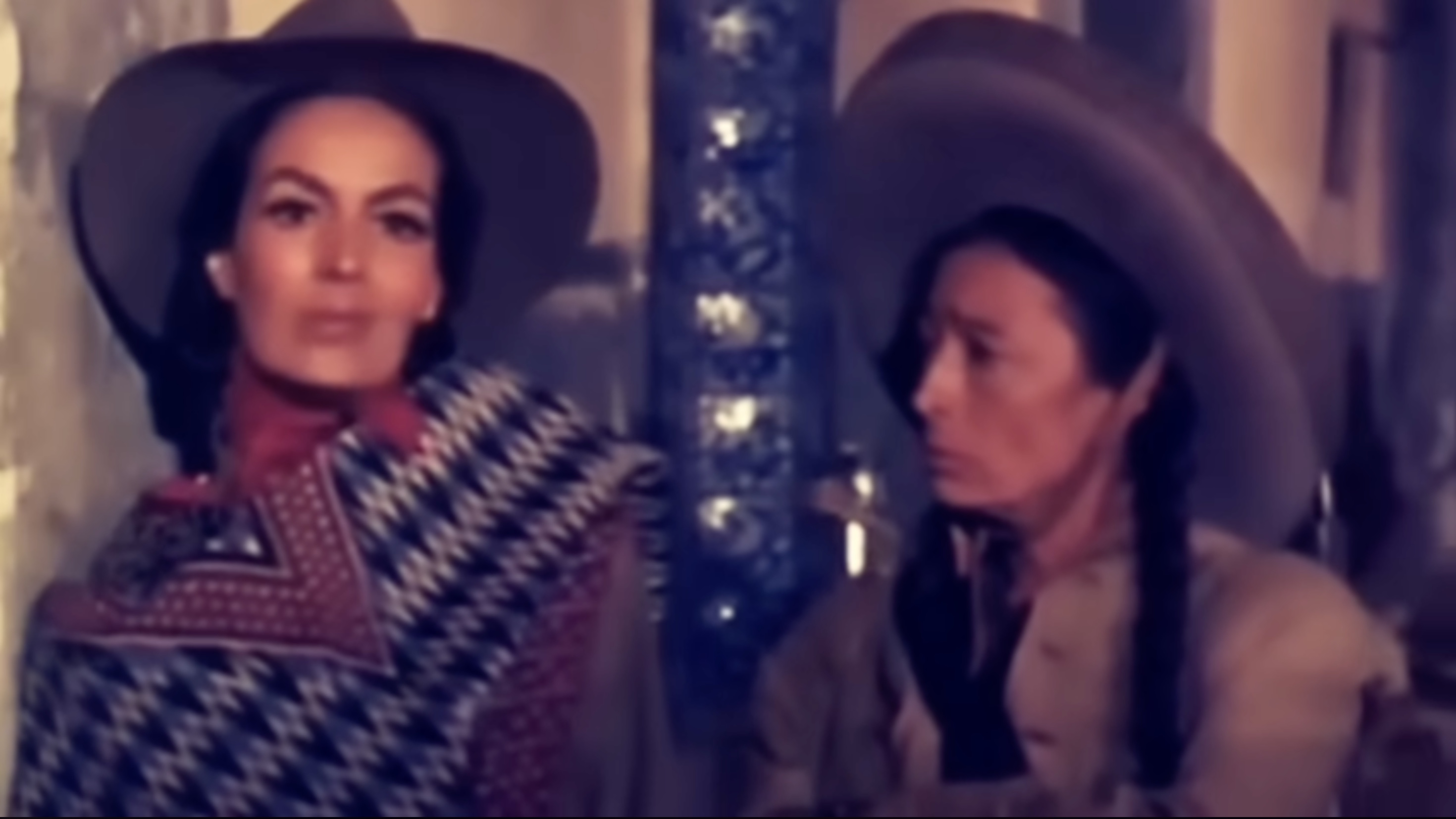 Still from La Cucaracha (1959) showing Cucaracha in a bold patterned poncho and wide-brimmed hat seated beside another woman in plainer clothing.