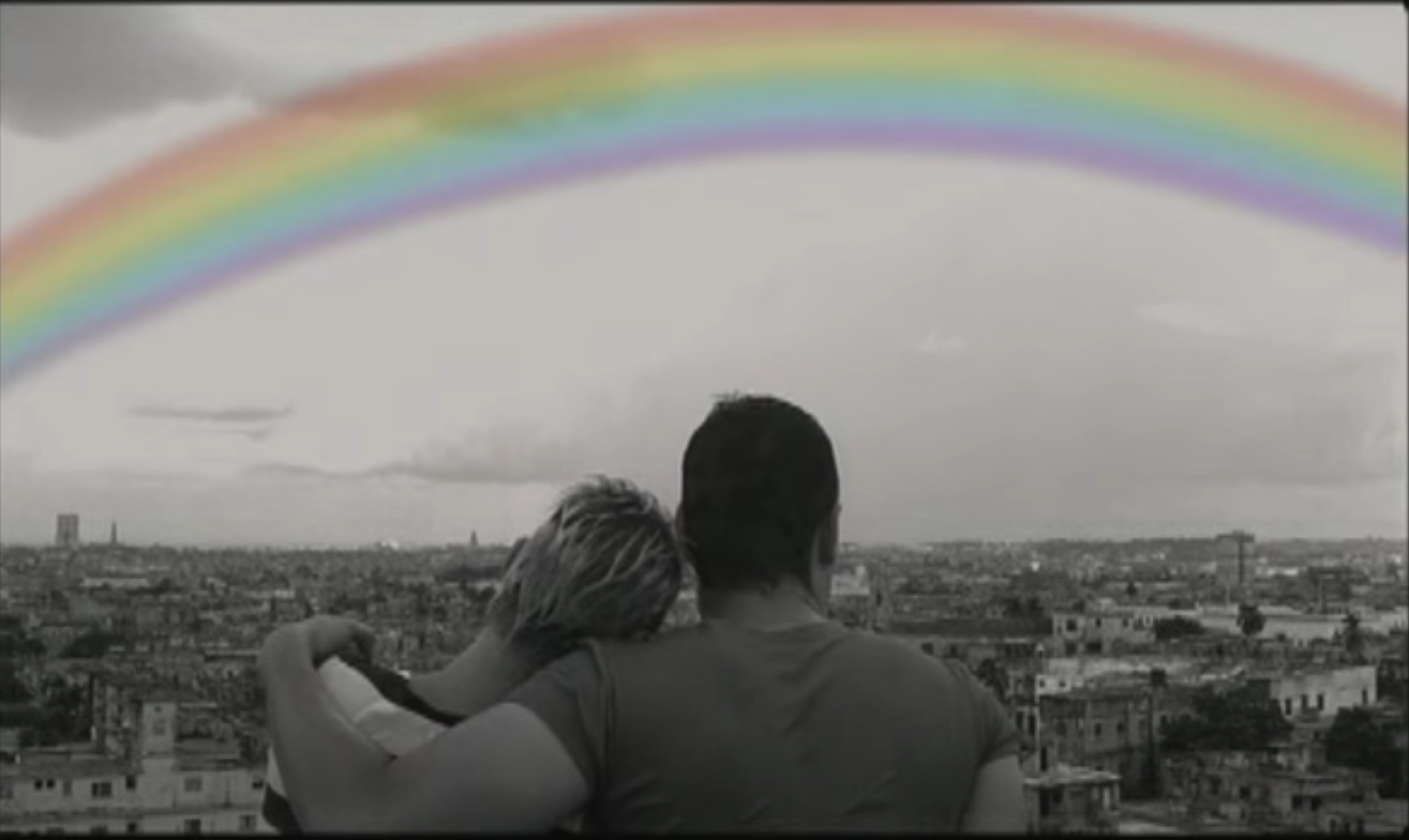 Black and white shot of César embracing Carla as they gaze at a colorful rainbow