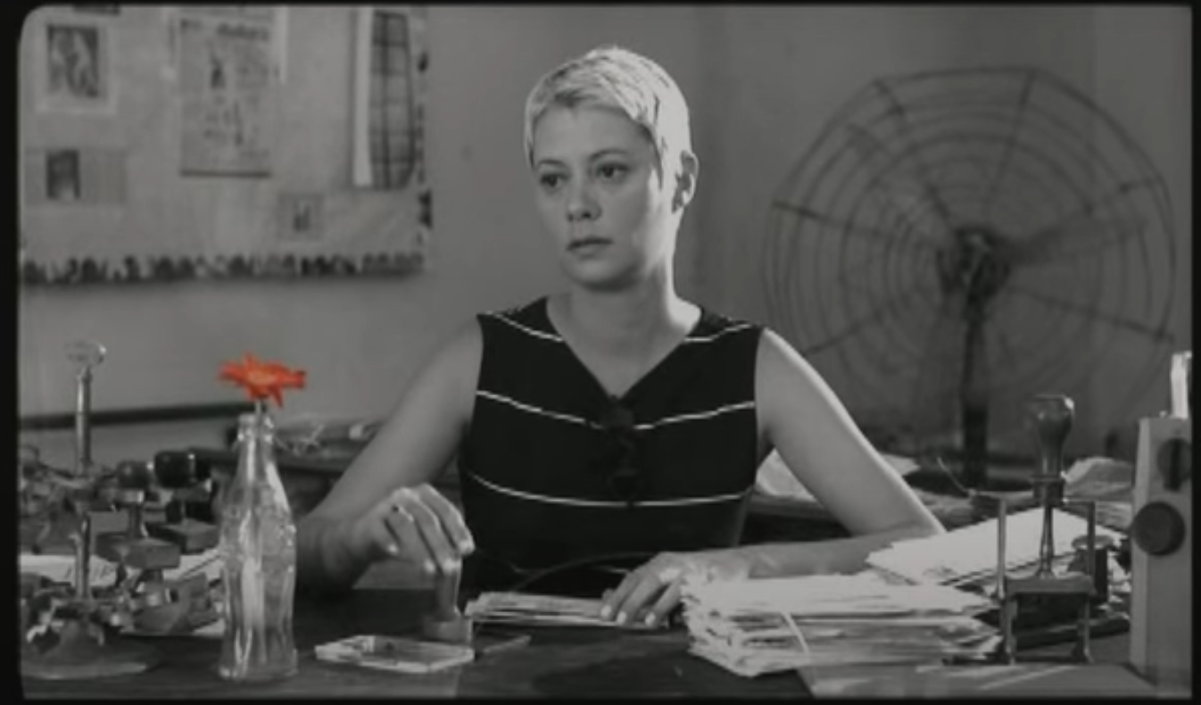 Black and white photo of Carla sitting in her post office desk with a red flower in a vase.