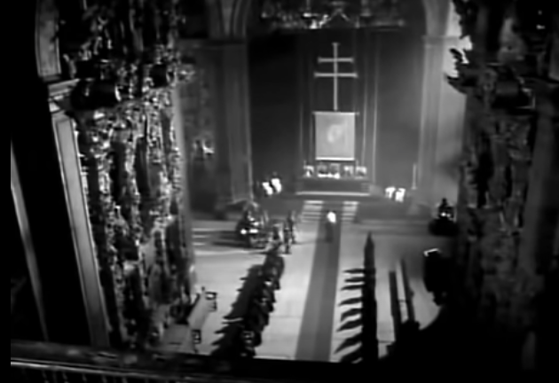 A extreme long shot taken from a high angle where we see the church, with Macario standing in front of the judges.