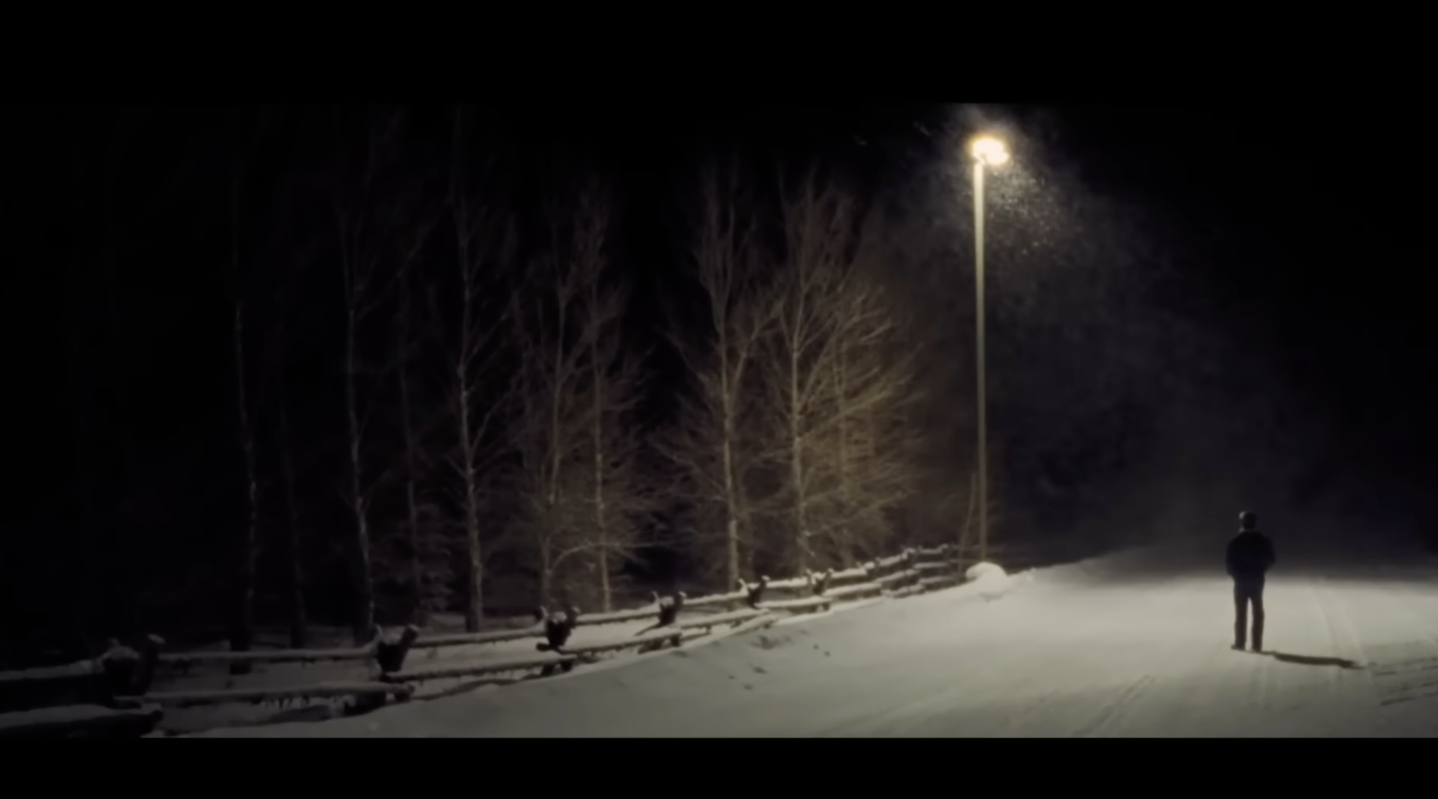 A lone figure stands on a snowy road at night under a single streetlamp as snow falls.