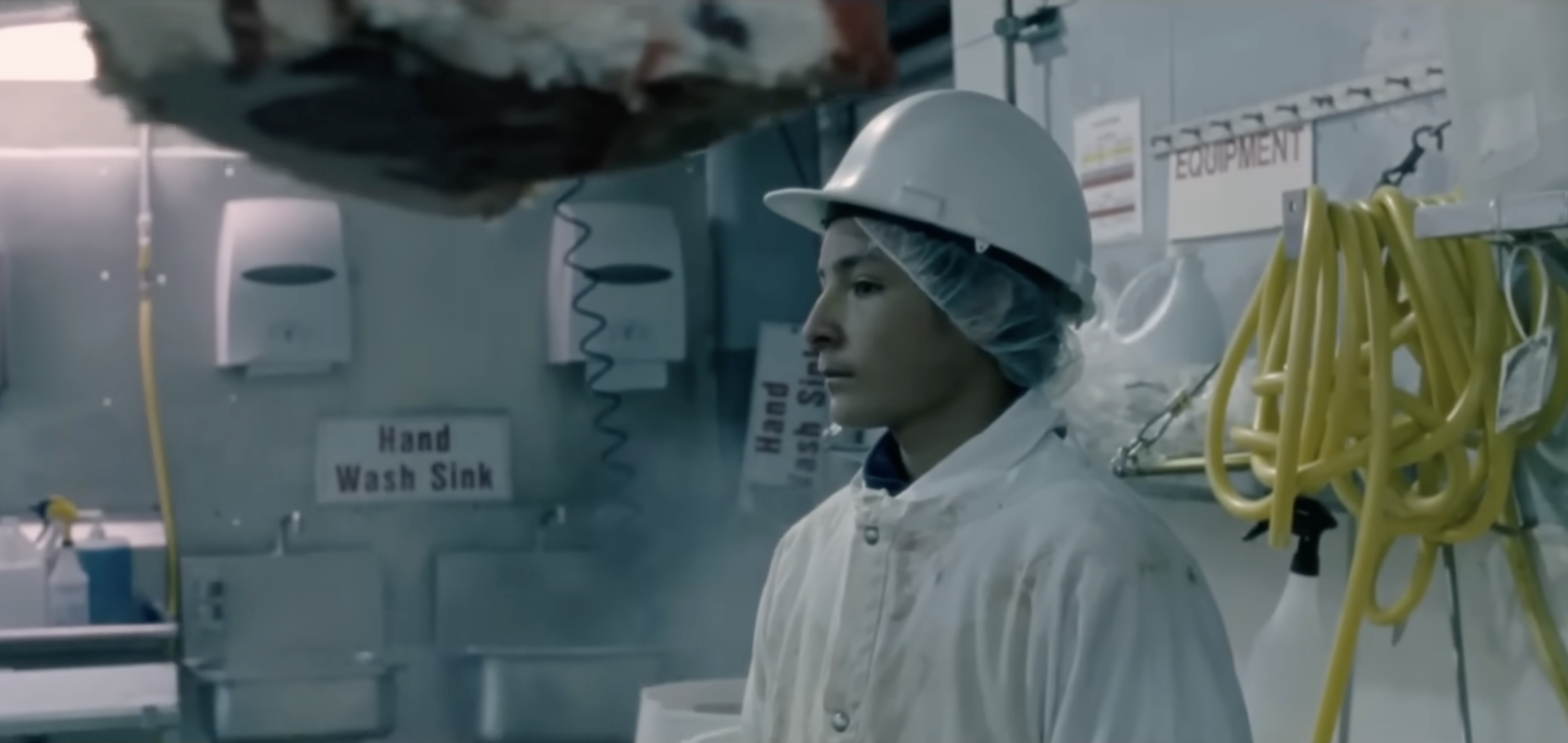 A teenage boy in a white coat, hairnet, and hard hat stands inside a meatpacking plant.