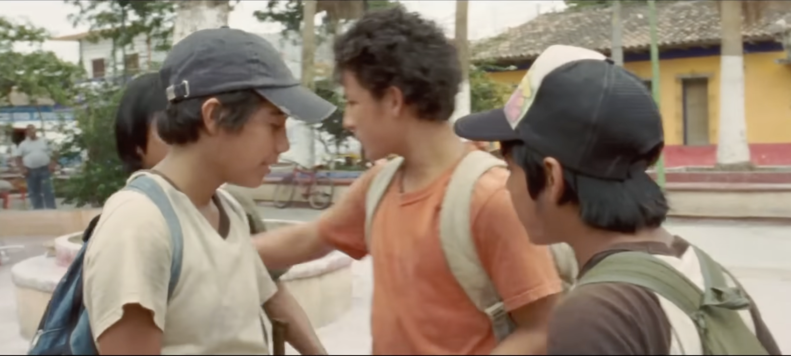 A boy in an orange shirt pushes another boy in a cap as a group of boys stands in a town plaza
