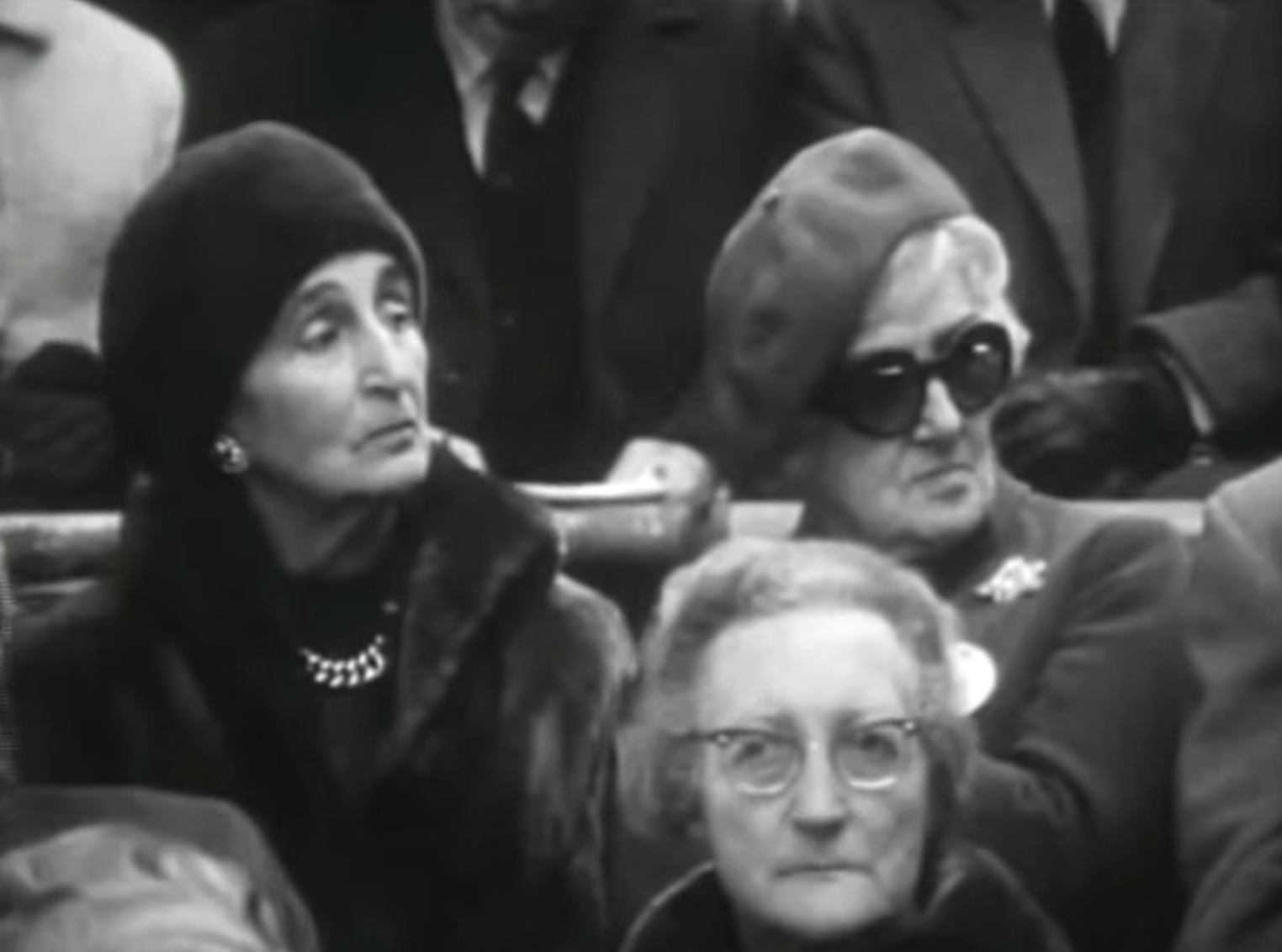 Wealthy upper-class women sit in spectatorship wearing thick coats, jewelry, formal berets, and oversized dark designer sunglasses.