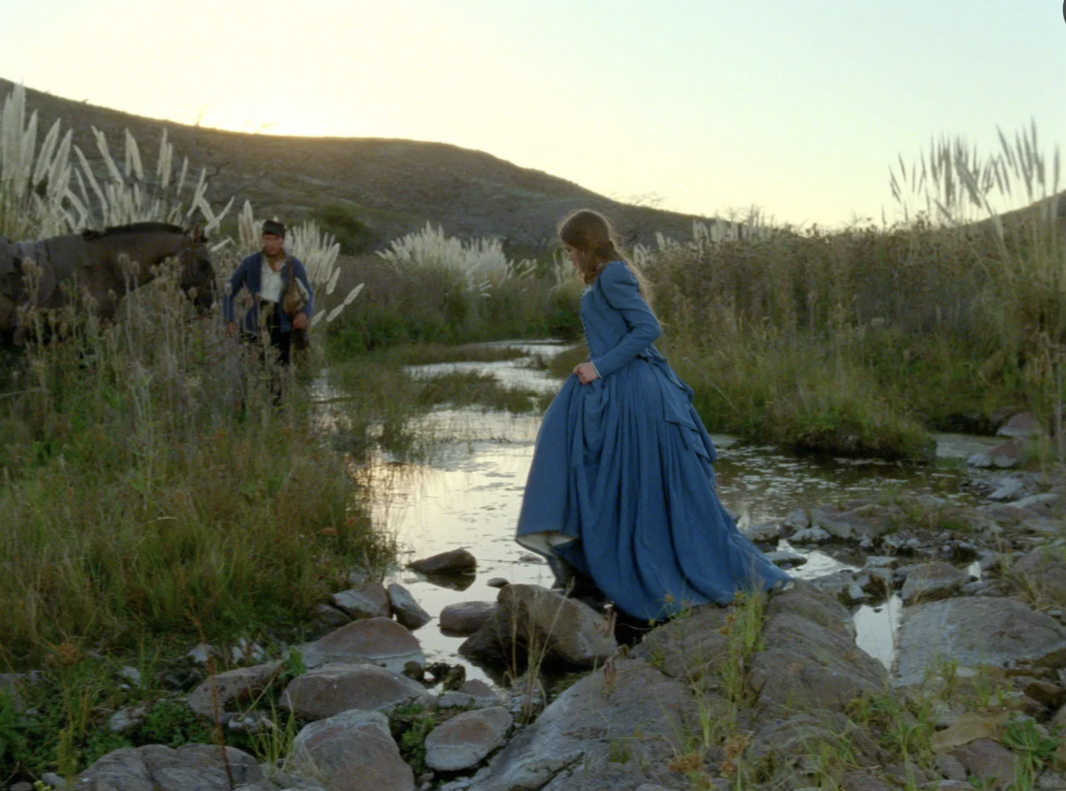 A woman uses some rocks to cross a lake in a 19th century gown. 