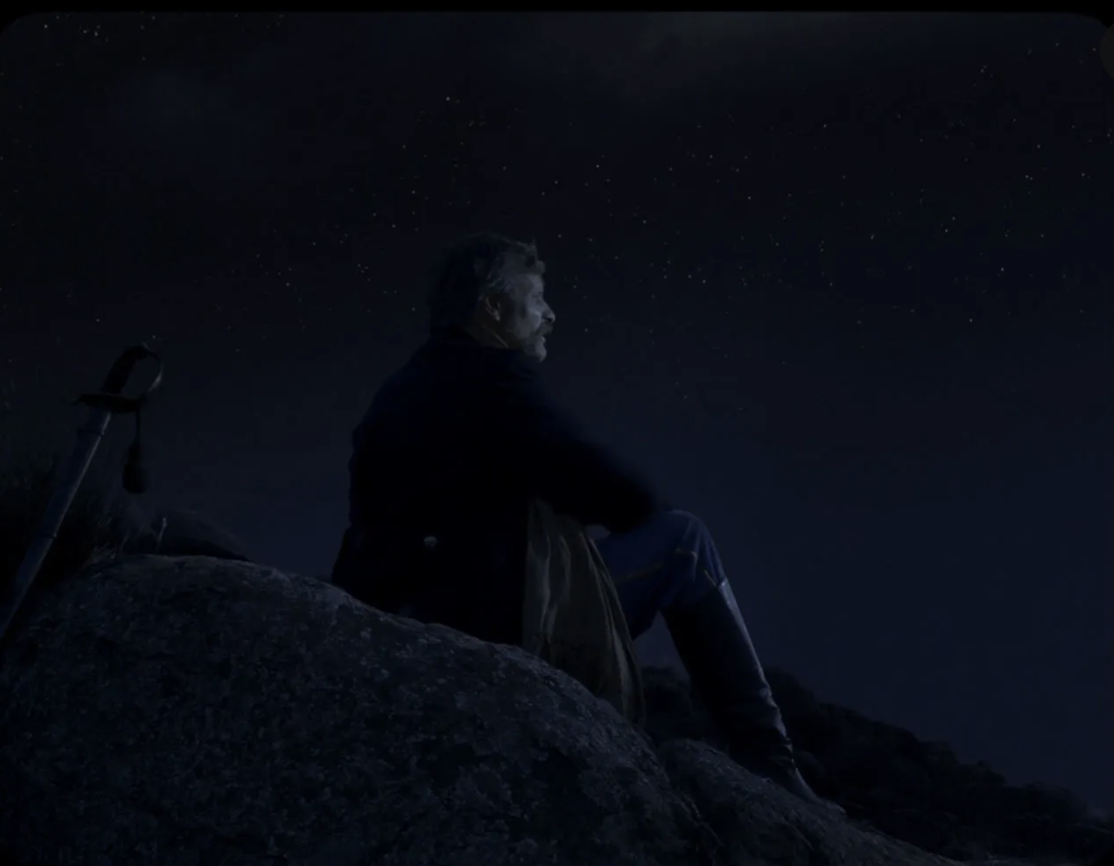 A man sits on a rock in the dark staring into the night sky. 