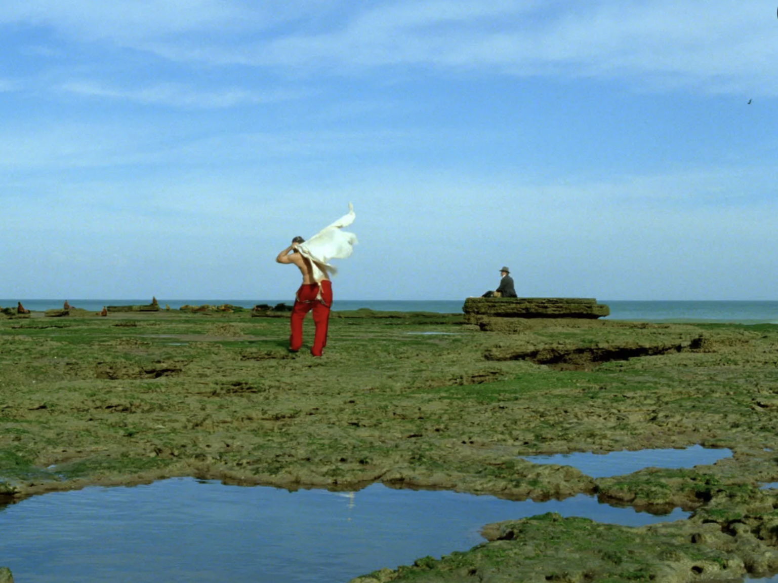 A man puts on his shirt as he walks to another man sitting on rock. The landscape around them is vast and the colors are very saturated.