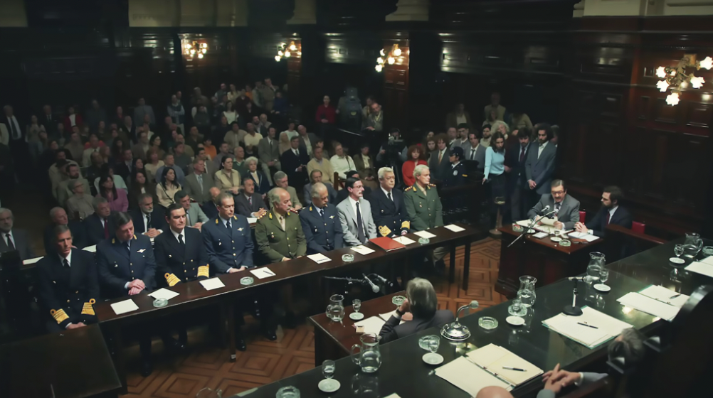 Courtoom lighting – low key with emphasis on prosecution, junta officials, and judge's table.