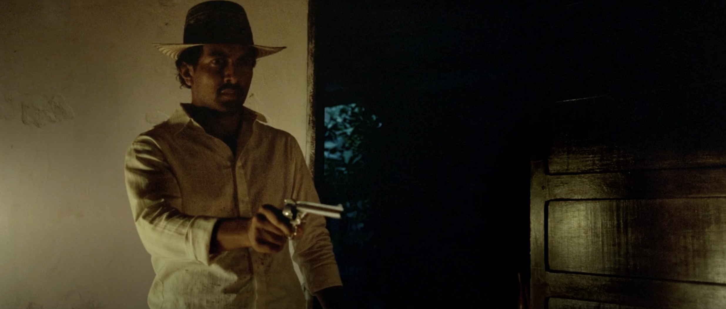 Rapayet holds a gun beside a dark doorway.
