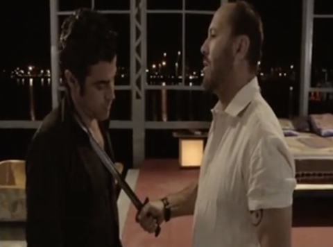 Dos hombres en un apartamento oscuro. Uno acaricia el pecho del otro con una espada.