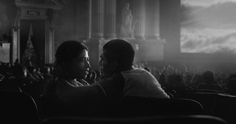 A black-and-white film still showing a young woman and man sitting beside each other in a movie theater. They turn toward one another while rows of seats and large decorative columns are visible in the background.