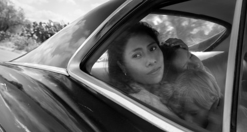 A black-and-white film still showing a woman seated inside a car with two children resting against her. She looks through the car window while reflections of clouds and the surrounding landscape appear across the glass.
