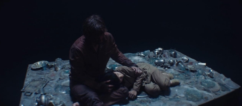 A man sitting on a platform covered in glass shards. A young girl is laying on his lap. The background is completely black.